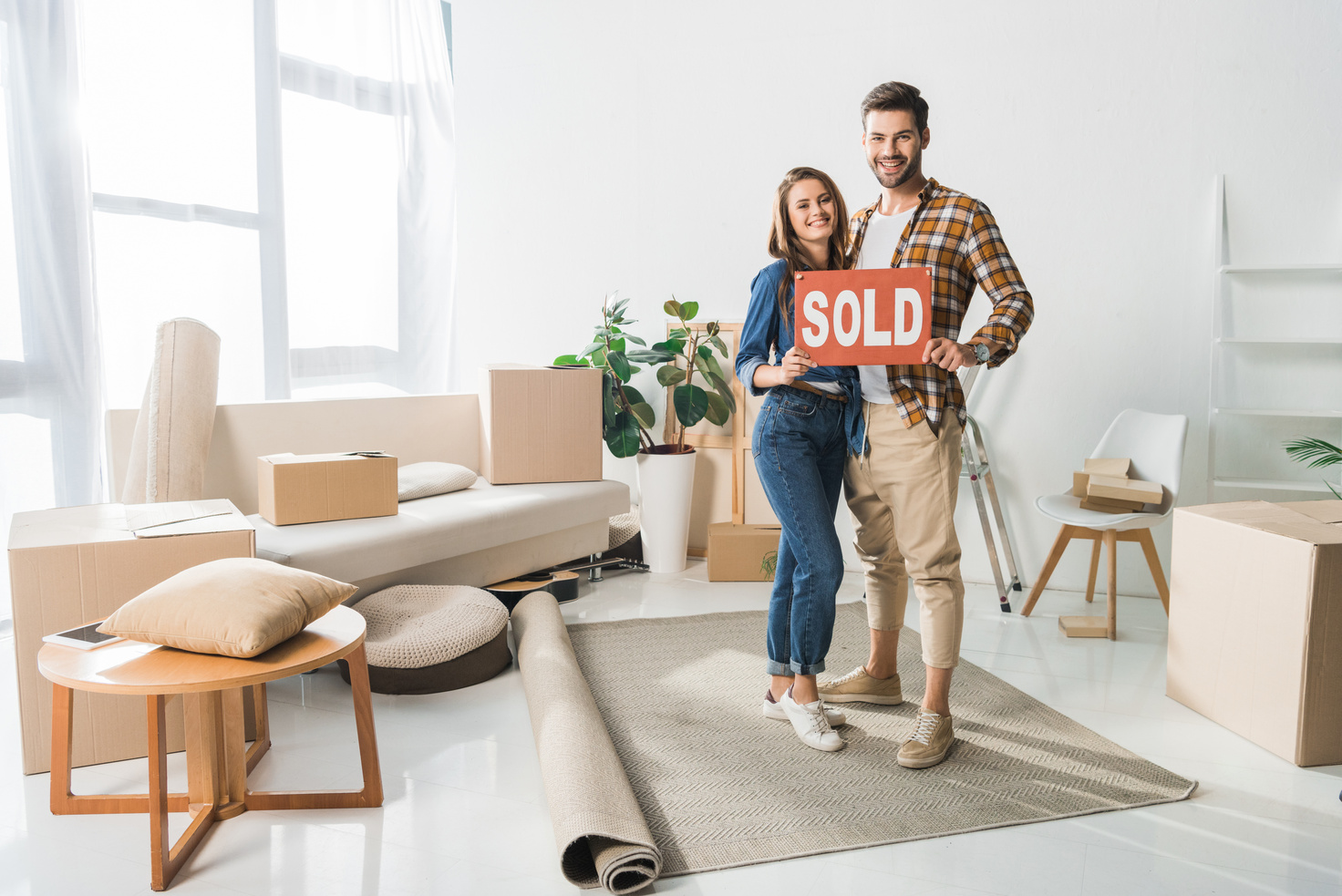 smiling couple holding sold red card at home with cardboard boxes